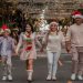 A joyful family walks down a decorated street wearing Christmas hats, symbolizing holiday spirit.
